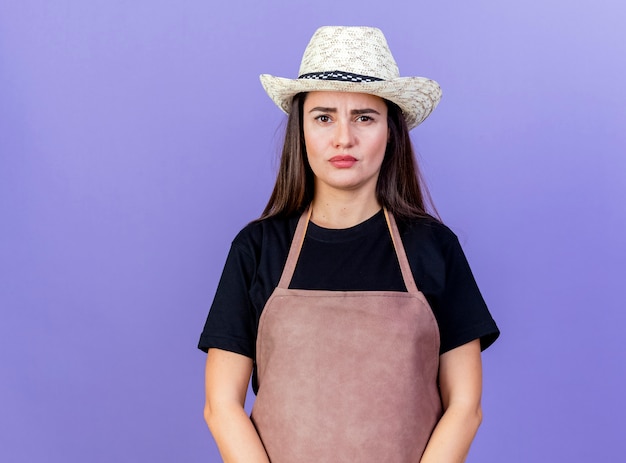 Kostenloses Foto unzufriedenes schönes gärtnermädchen in der uniform, die den auf blau isolierten gartenhut trägt