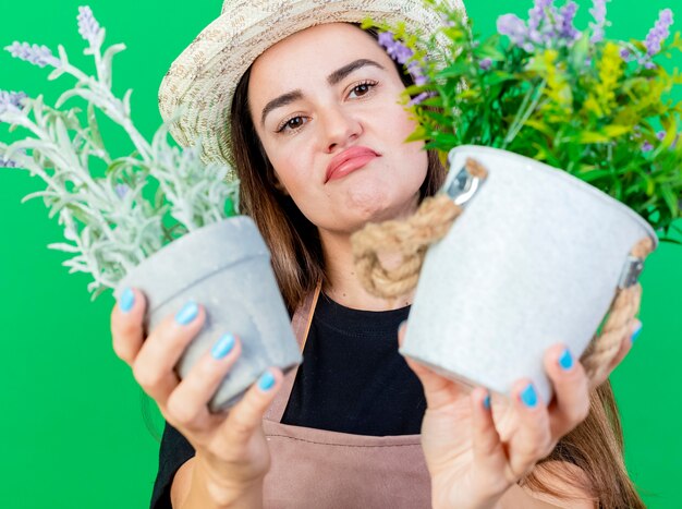 Unzufriedenes schönes Gärtnermädchen in der Uniform, die Gartenhut trägt, der Blume im Blumentopf heraushält, der auf Grün lokalisiert wird