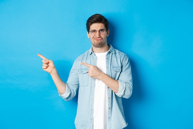 Unzufriedenes und skeptisches männliches Model mit Brille, das mit den Fingern auf etwas Schlechtes zeigt, schreckliche Werbung zeigt und auf blauem Hintergrund steht.