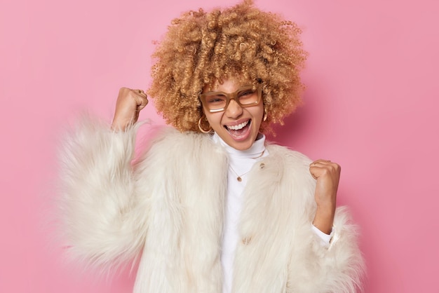 Kostenloses Foto upbeat energische junge frau mit lockigem haar macht fauststoß feiert erfolg schreit vor freude trägt transparente brille und wintermantel isoliert über rosa hintergrund ja, ich habe es endlich geschafft