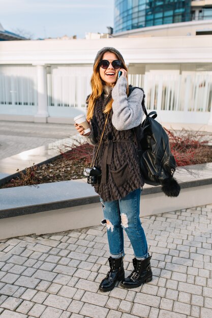 Urban modische junge Frau in der kühlen Sonnenbrille, im warmen Winterpullover, in den stilvollen Jeans, die mit Rucksack in der Stadt reisen. Fröhliche Stimmung, telefonieren, Kaffee zum Mitnehmen, sonniger kalter Tag.