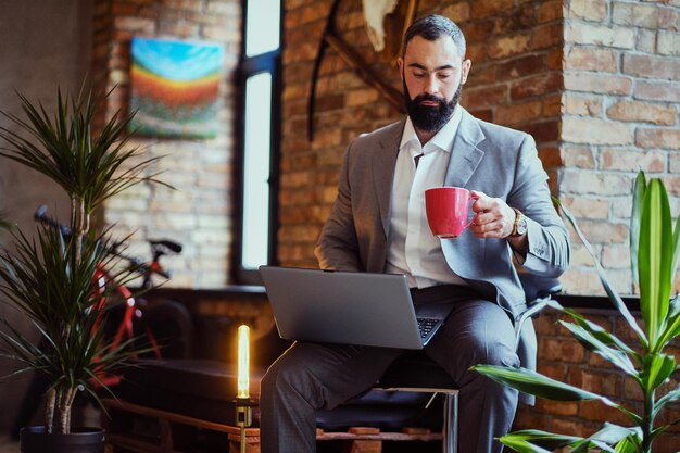 Urbaner bärtiger Mann trinkt Kaffee und benutzt einen Laptop in einem Raum mit Loft-Interieur.