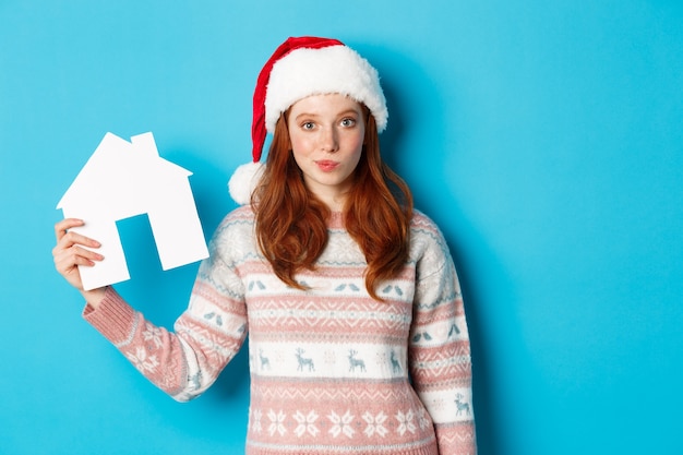 Urlaubsangebote und Immobilienkonzept. Süße rothaarige Frau in Nikolausmütze und Pullover mit Papierhausmodell, Wohnungsangebot, stehend auf blauem Hintergrund.