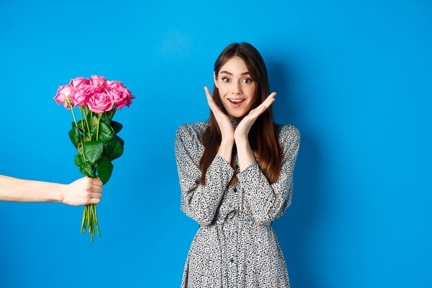 Valentinstag-Konzept. Aufgeregte und glückliche junge Frau, die erstaunt in die Kamera schaut, während die Hand mit Blumenstrauß die Hand ausstreckt und ein romantisches Geschenk erhält, blauer Hintergrund