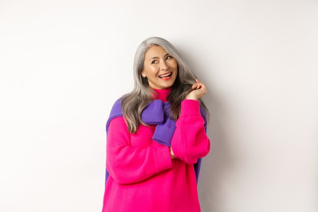 Valentinstag-Konzept. Schöne asiatische Seniorin, die kokett lacht, mit Haarsträhnen spielt und die obere linke Ecke flirty betrachtet, auf weißem Hintergrund stehend