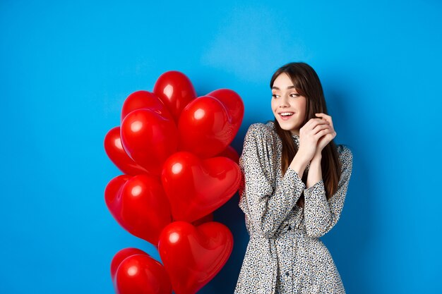 Valentinstag. Schönes romantisches Mädchen, das vom Datum träumt, in der Nähe von schönen Herzballons steht und lächelt, blauer Hintergrund.