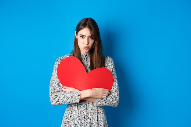 Valentinstag trauriges und düsteres kaukasisches Mädchen verzieht die Lippen und sieht enttäuscht aus, hält großes rotes...