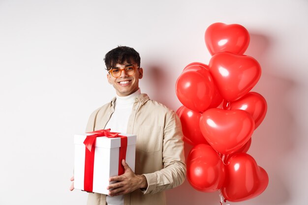 Valentinstag und Romantik-Konzept. Der verliebte Mann bereitet ein Überraschungsgeschenk für den Liebhaber vor, hält das Geschenk in der Schachtel und steht in der Nähe der Geste der roten Herzen, weißer Hintergrund.