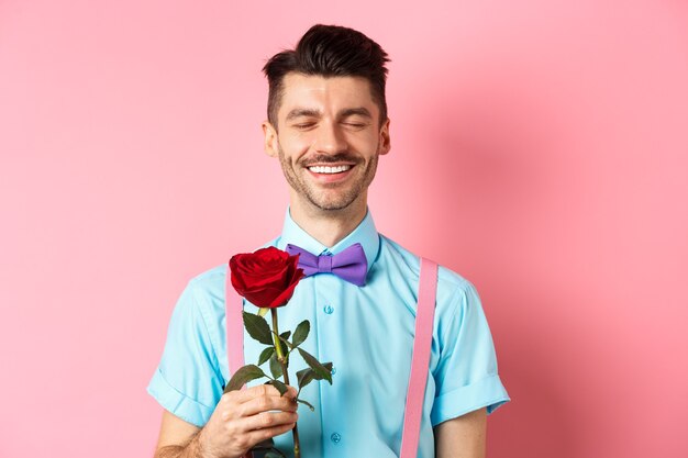 Valentinstag und Romantik-Konzept. Romantischer Mann mit roter Rose, der mit Liebhaber verabredet ist und in schicker Fliege auf rosa Hintergrund steht.