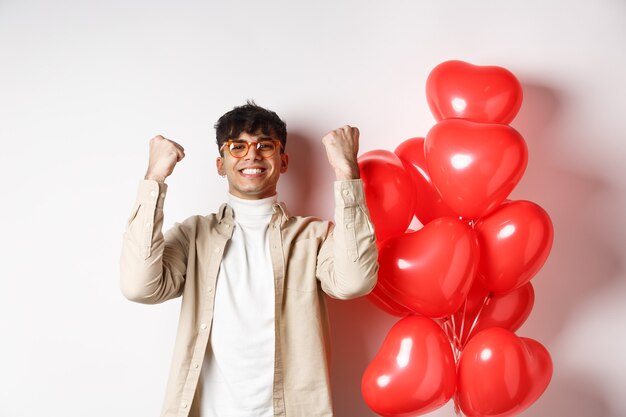 Valentinstag. Zufriedener junger Mann sagt ja, triumphiert und feiert am Liebhaberdatum, macht Faustpumpe und lächelt zufrieden, steht in der Nähe von Herzballons auf weißem Hintergrund.