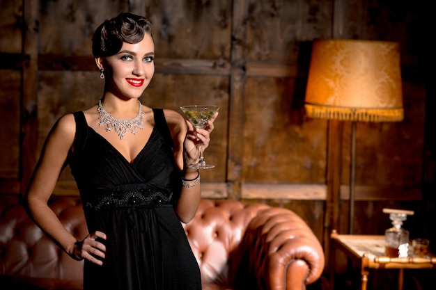 Kostenloses Foto vamp-dame mit roten lippen posiert mit einem glas cocktail im restaurant. elegante reiche dame im schwarzen kleid, die mit ihrer hand auf der hüfte posiert. vermögenskonzept.