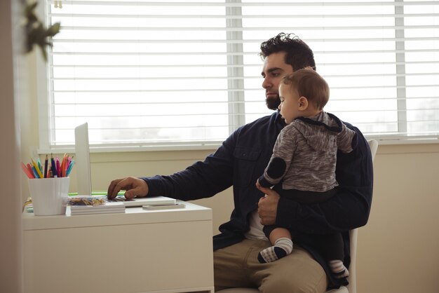 Vater hält sein Baby, während er Laptop am Schreibtisch benutzt