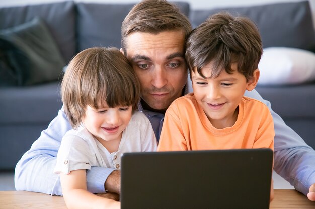 Vater mittleren Alters umarmt Söhne und schaut sich einen Film auf dem Laptop an. Glückliche kleine Jungen, die mit Vater am Tisch sitzen, Bildschirm betrachten und lächeln. Konzept für Vaterschaft, Kindheit und digitale Technologie
