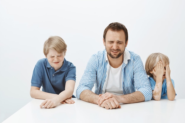 Vater und Söhne sind verärgert, während Mama bei der Arbeit ist. Porträt der hungrigen unzufriedenen europäischen Familie der männlichen Jungen und des Vaters, die am Tisch sitzen und weinen