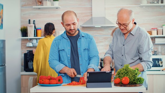 Vater und Sohn kochen Gemüse zum Abendessen mit Online-Rezept auf dem PC-Computer in der heimischen Küche. Männer, die beim Zubereiten der Mahlzeit ein digitales Tablet verwenden. Große Familie gemütliches erholsames Wochenende.