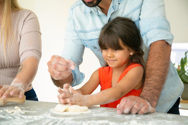 Vater zeigt Tochter, wie man Teig auf Küchentisch mit Mehl unordentlich macht. Junges Paar und ihr Mädchen backen Brötchen oder Kuchen zusammen. Familienkochkonzept