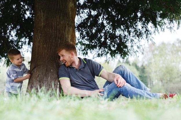 Vati und Sohn spielen Verstecken, sitzen unter dem Baum im Park