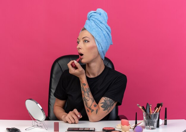 Verängstigt aussehende junge schöne Mädchen sitzt am Tisch mit Make-up-Tools, die Haare in ein Handtuch gewickelt haben und Lippenstift einzeln auf rosafarbenem Hintergrund auftragen