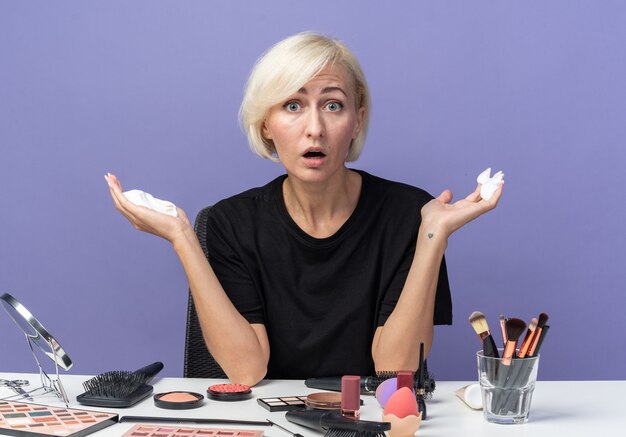 Verängstigt in die Kamera schaut junges schönes Mädchen sitzt am Tisch mit Make-up-Tools, die Haarcreme isoliert auf blauem Hintergrund halten