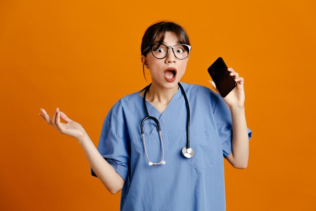 Verängstigte sich ausbreitende Hand, die das Telefon hält Junge Ärztin mit einheitlichem Fith-Stethoskop isoliert auf orangefarbenem Hintergrund