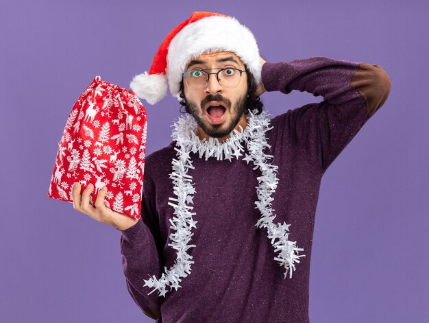 verängstigter junger gutaussehender Kerl, der Weihnachtsmütze mit Girlande am Hals trägt und Weihnachtstasche hält, die Hand hinter den Kopf legt, isoliert auf blauer Wand?