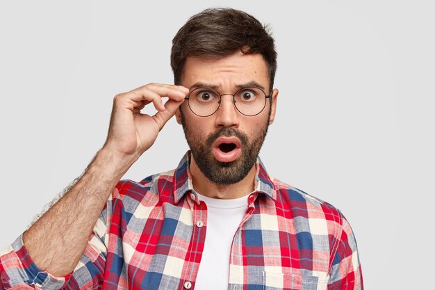 Verängstigter, verwirrter Mann mit verängstigtem Gesichtsausdruck, starrt durch die Brille, öffnet den Mund weit und hält die Hand am Rand der Brille