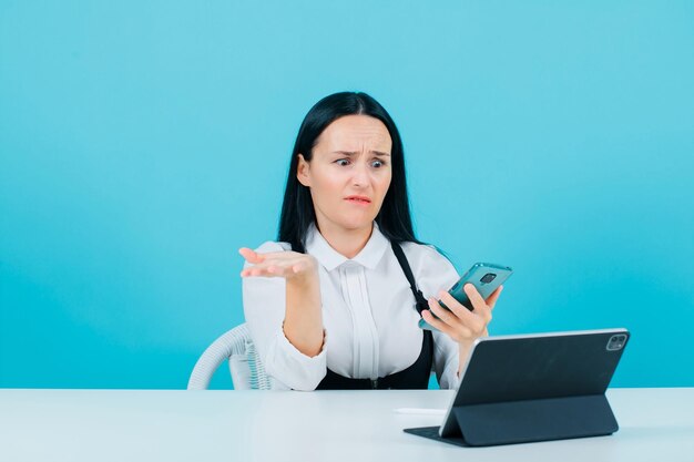 Verängstigtes Blogger-Mädchen schaut auf ihren Telefonbildschirm, indem sie vor einem Tablet auf blauem Hintergrund sitzt