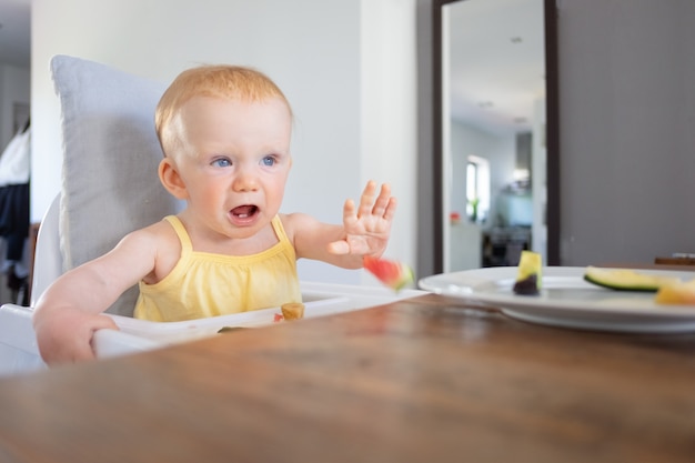 Verärgert süßes Baby, das im Hochstuhl sitzt und Scheiben der Früchte wirft. Erstes Konzept für feste Lebensmittel oder Kinderbetreuung