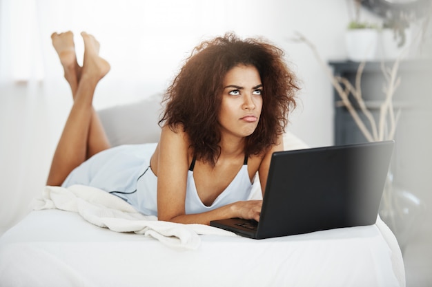 Verärgert traurige schöne afrikanische Frau in Nachtwäsche liegend mit Laptop auf Bett zu Hause denken.