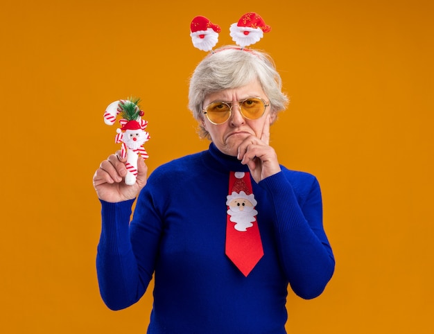 Verärgerte ältere Frau in Sonnenbrille mit Santa Stirnband und Santa Krawatte legt Hand auf Kinn und hält Zuckerstange isoliert auf orange Hintergrund mit Kopienraum