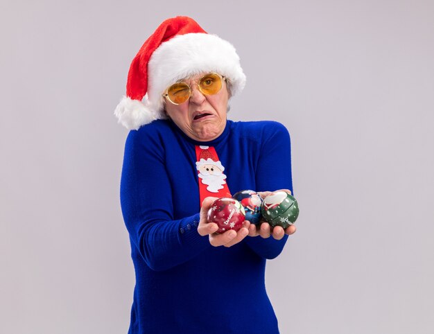 Verärgerte ältere Frau in Sonnenbrille mit Weihnachtsmütze und Weihnachtskrawatte, die Glaskugelverzierungen hält