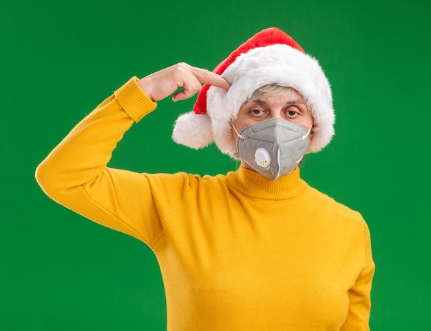 Verärgerte ältere Frau mit Weihnachtsmütze, die medizinische Maske trägt, setzt Finger auf Tempel lokalisiert auf grünem Hintergrund mit Kopienraum