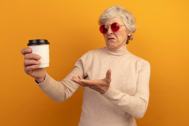 Verärgerte alte Frau mit cremigem Rollkragenpullover und Sonnenbrille, die eine Plastiktasse Kaffee hält und mit der Hand darauf zeigt