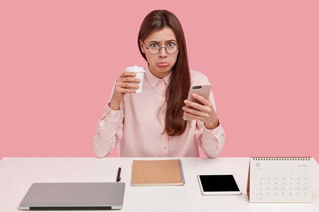 Verärgerte brünette weibliche Büro-Perfektionistin hat einen unglücklichen Ausdruck, trinkt Kaffee zum Mitnehmen, hält ein Handy in der Hand und trägt eine runde Brille