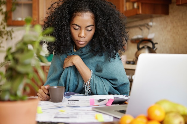 Verärgerte dunkelhäutige Hausfrau mit Afro-Frisur, die Kaffee trinkt, während sie spät in der Nacht das Haushaltsbudget verwaltet