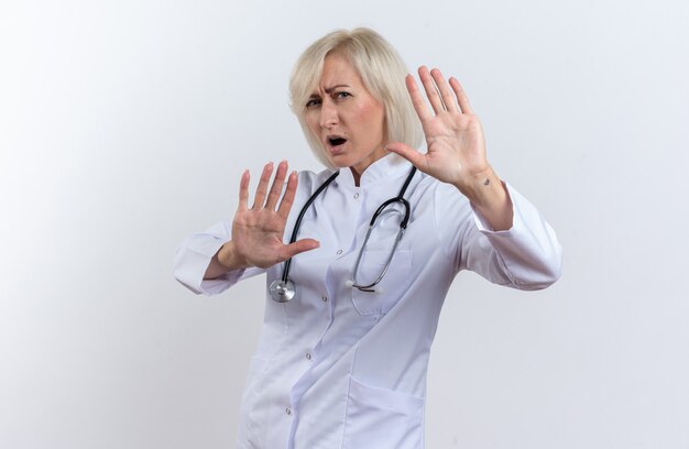 Verärgerte erwachsene slawische Ärztin in medizinischer Robe mit Stethoskop, die mit erhobenen Händen auf weißem Hintergrund mit Kopienraum steht