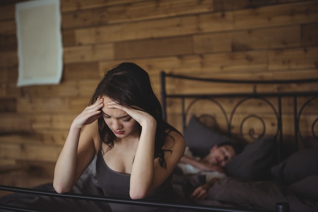Verärgerte Frau, die auf einem Bett sitzt, während Mann im Hintergrund schläft
