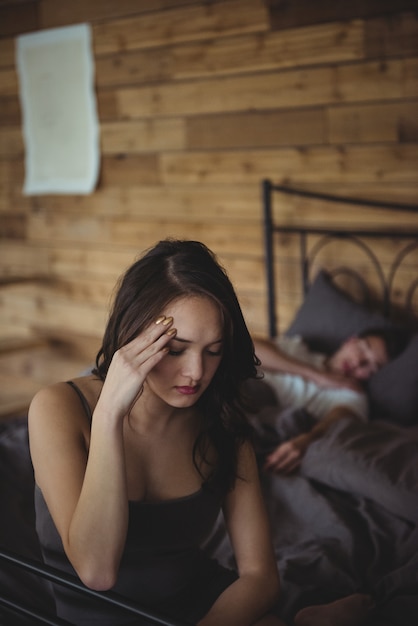 Verärgerte Frau, die auf einem Bett sitzt, während Mann im Hintergrund schläft