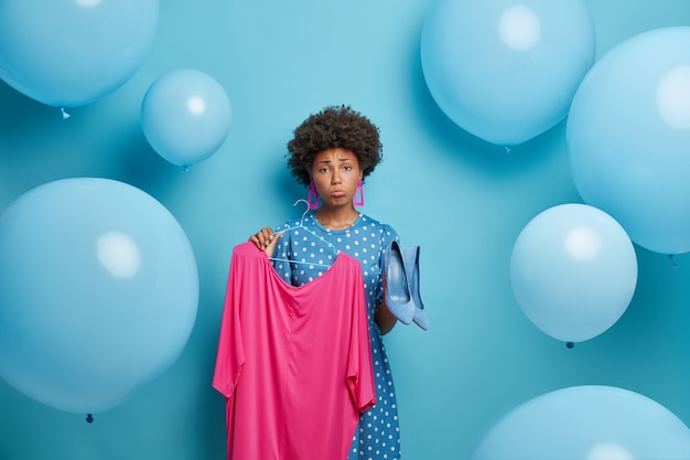 Verärgerte Frau hat ein Problem damit, was sie anziehen soll, hält rosa Kleid auf Kleiderbügel und blaue Schuhe mit hohen Absätzen, traurige Kleidungsstücke passen nicht zusammen, wählt Outfit für besondere Anlässe, drückt negative Emotionen aus