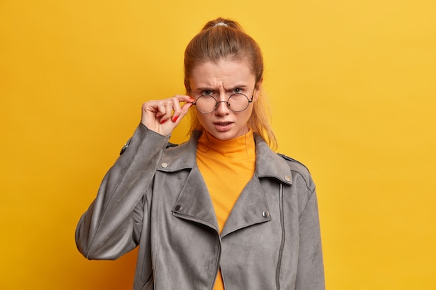 Verärgerte Frau schaut gewissenhaft durch transparente Brille, stimmt mit etwas nicht überein, in graue Jacke gekleidet, posiert