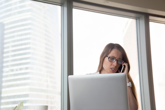 Verärgerte geschäftsfrau, die auf mobile am arbeitsplatz, argumentierend durch zelle spricht