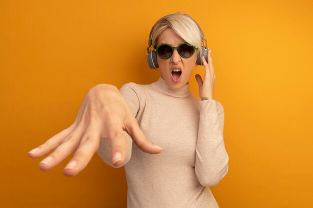 Verärgerte junge blonde Frau mit Sonnenbrille und Kopfhörern, die Kopfhörer berühren