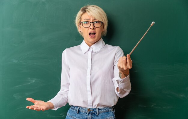 Verärgerte junge blonde Lehrerin mit Brille im Klassenzimmer, die vor der Tafel steht und einen Zeiger hält, der nach vorne schaut und leere Hand zeigt