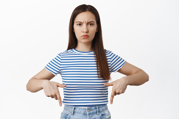 Verärgerte junge Frau, die mit dem Finger nach unten zeigt, die Stirn runzelt und wütend in die Kamera schaut, sich über Banner beschwert, schlechte Werbung, etwas fürchterlich beurteilt, weißer Hintergrund.