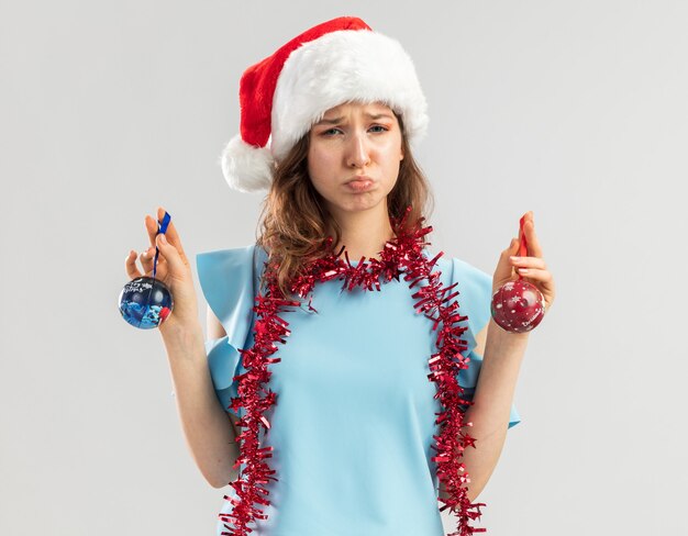 Verärgerte junge Frau im blauen Oberteil und im Weihnachtsmannhut mit Lametta um ihren Hals, der Weihnachtskugeln hält, die mit traurigem Ausdruck schauen