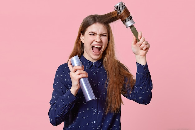 Verärgerte junge Frau kämmt sich die Haare, ist mit Shampoo unzufrieden, hält Haarbürste und Haarspray in der Hand und schaut mit gereiztem Gesichtsausdruck in die Kamera