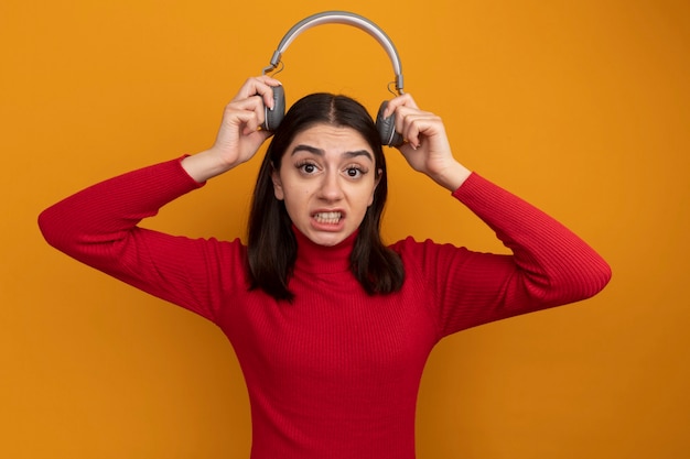 Verärgerte junge hübsche Frau, die Kopfhörer über dem Kopf hält und nach vorne auf orangefarbene Wand schaut?