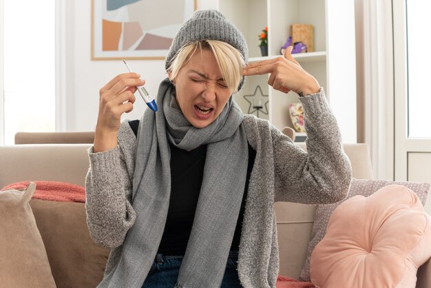 Verärgerte junge kranke slawische Frau mit Schal um den Hals, die eine Wintermütze trägt, die Hand an ihrem Tempel hält, das Waffenschild gestikuliert und das Thermometer auf der Couch im Wohnzimmer hält