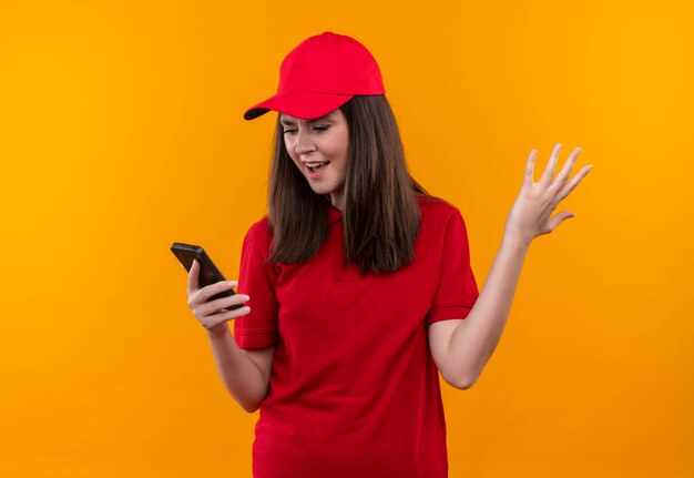 Verärgerte junge Lieferfrau, die rotes T-Shirt in der roten Kappe hält, die ein Telefon auf isolierter gelber Wand hält