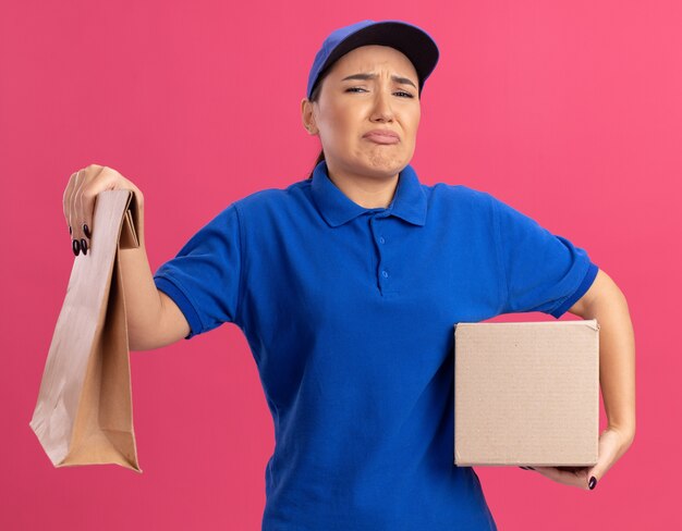 Verärgerte junge Lieferfrau in blauer Uniform und Kappe, die Papierpaket und Pappkarton hält, der vorne mit traurigem Ausdruck steht, der über rosa Wand steht